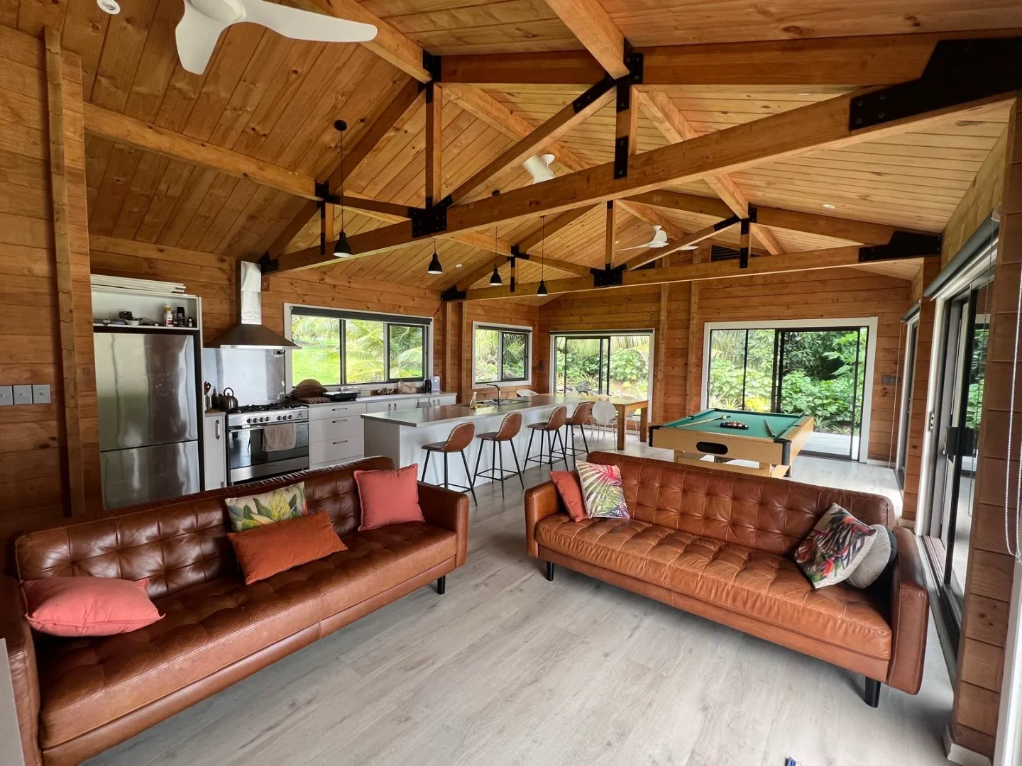Inside Rotuma Stay — open-plan living area with leather sofas, kitchen, pool table, and vaulted timber ceiling